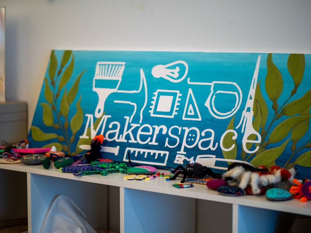 Horizontal canvas painted blue with sea kelp and the makerspace logo leaned up against a wall and shelves.
