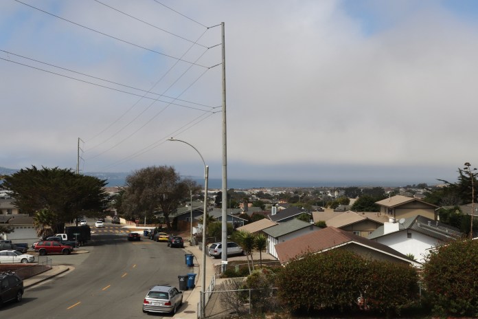 Landscape photo of suburban area. Top of houses, telephone poles, street lights, and cars in the foreground. Clouds and the ocean are visible in the distance.
