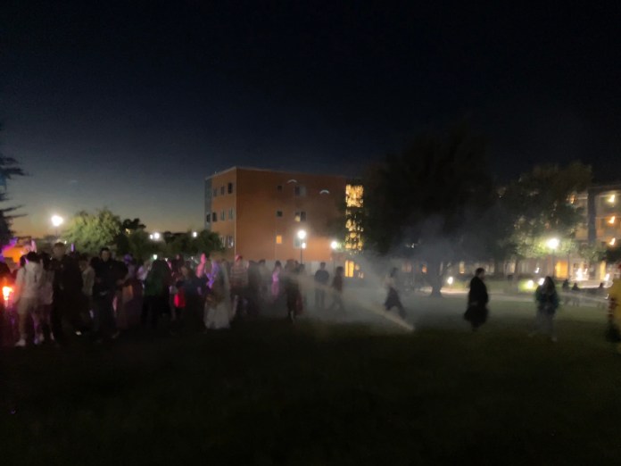 Dimly lit photo of people standing outside on the grass getting sprayed by sprinklers.