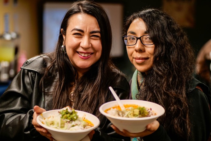 Two people wearing dark clothing holding up bowls of pozole, smiling for a photo.