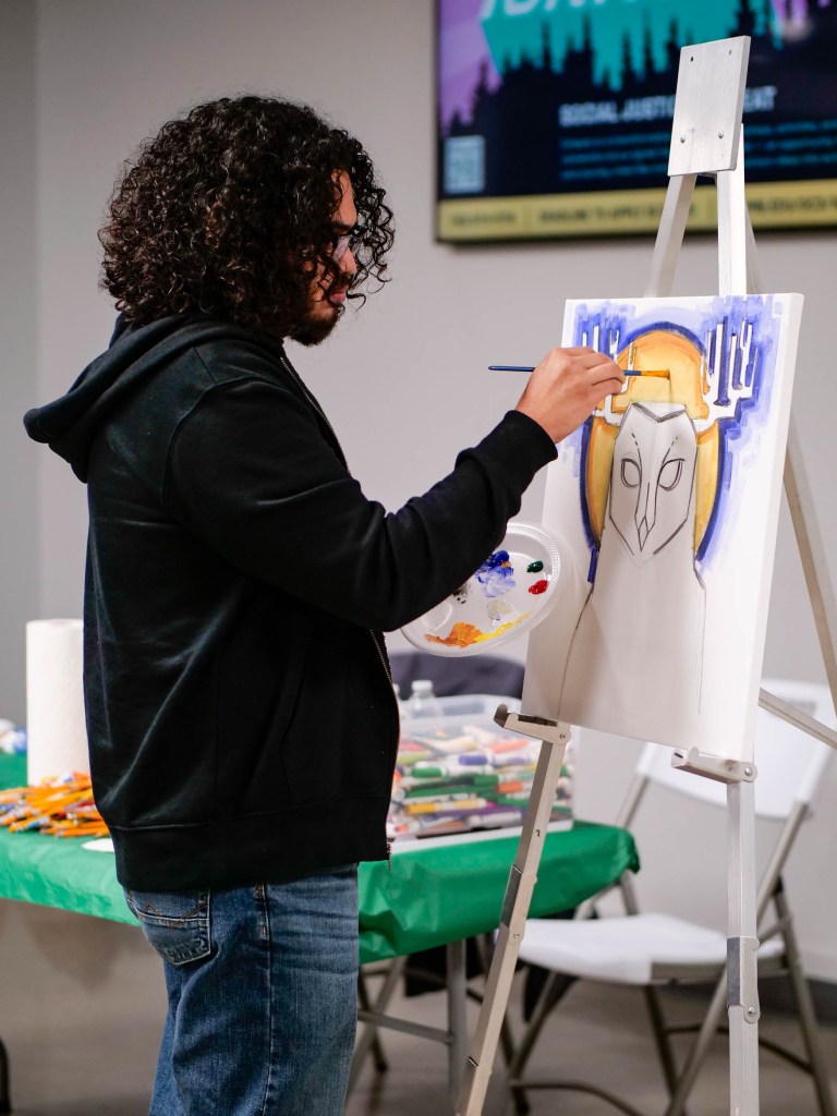 Person with dark curly hair wearing a black hoodie painting at a standing easel.