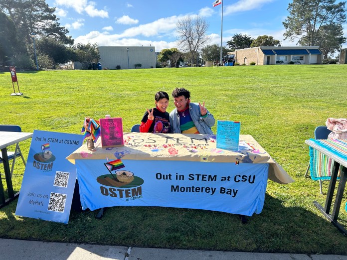 Two people sitting at a tabling event. Tablecloth reads 'Out in STEM at CSU Monterey Bay.