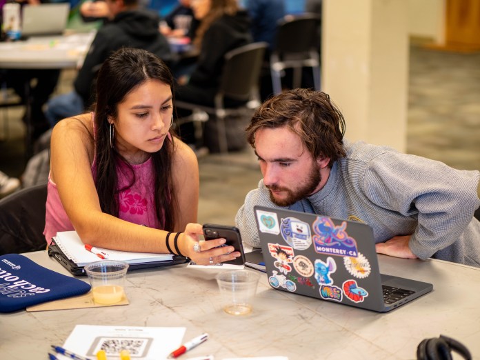 Two people sitting at a table. One with a laptop, the other showing something on their phone.
