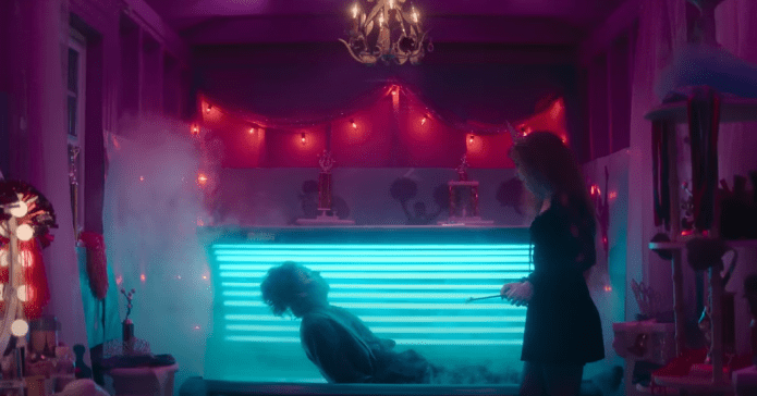 Person sitting up out of a tanning bed. Another person standing, watching. Blue and pink lighting.