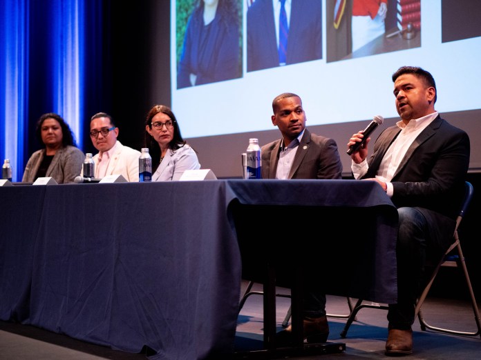 Five panelists sitting at a table on stage in front of a projection.