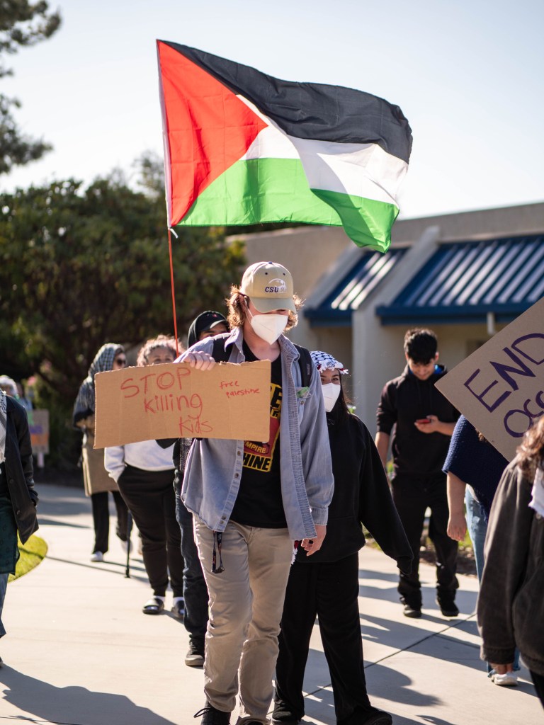 Protester holding sign that says Stop killing kids. Palestine flag behind him.