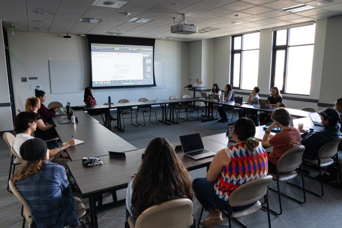 Photo of people sitting in a big desk circle with a projector in the middle of the room.