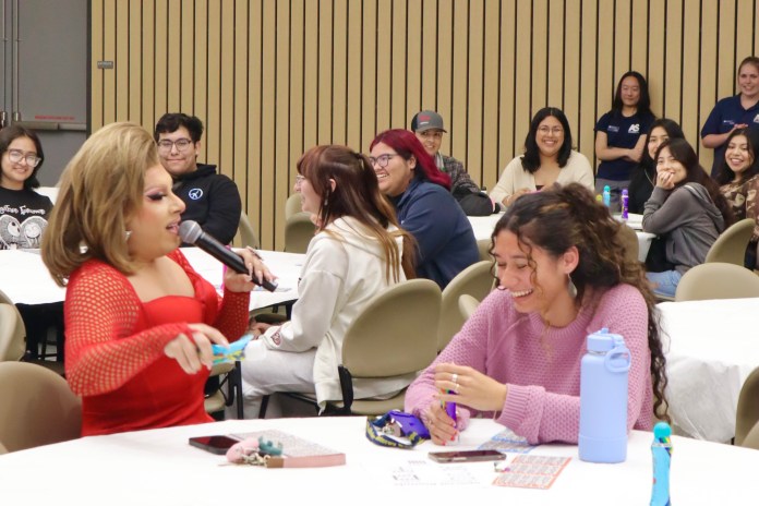 picture of a drag star speaking into a microphone with students in the background